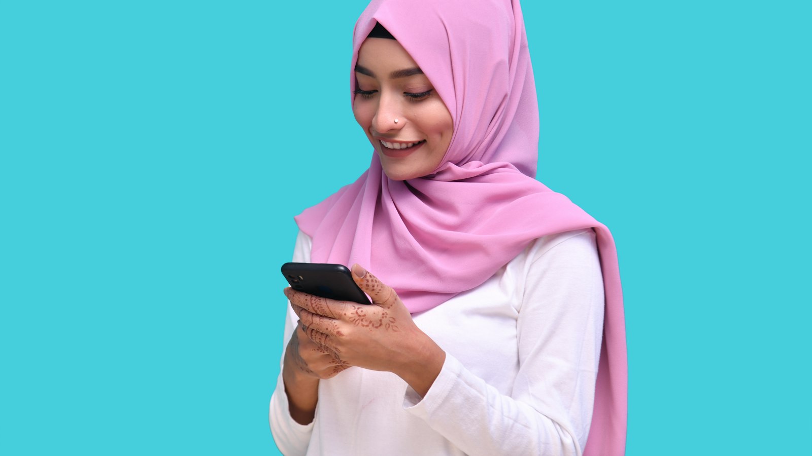 A smiling woman in a pink hijab with mehndi on her hands reading a message on her phone